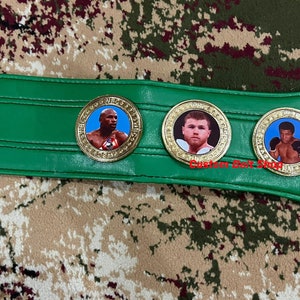 May include: A green boxing championship belt with three gold medallions featuring images of boxers. The medallions have a white background and the boxers are wearing boxing gloves.
