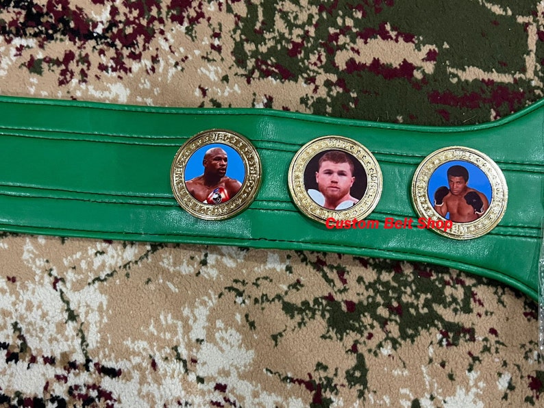 May include: A green boxing championship belt with three gold medallions featuring images of boxers. The medallions have a white background and the boxers are wearing boxing gloves.