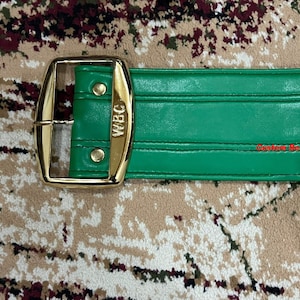 May include: A green leather boxing championship belt with gold accents. The belt has a gold buckle and a gold plate with the letters "WBC" on it. The belt also has a gold medallion with a picture of a boxer on it.