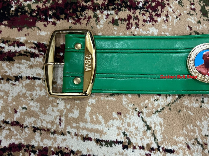 May include: A green leather boxing championship belt with gold accents. The belt has a gold buckle and a gold plate with the letters "WBC" on it. The belt also has a gold medallion with a picture of a boxer on it.