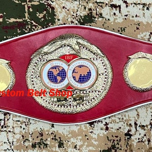 Op de afbeelding: Een rode leren bokskampioensgordel met gouden accenten en een witte rand. De riem heeft twee ronde platen met een wereldbeeld en de tekst "World Boxing Union" en "Custom Belt Shop".