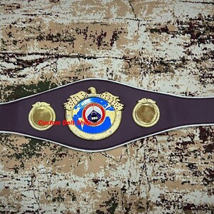 Op de afbeelding: Een gouden en blauwe bokskampioensgordel met het logo van de World Boxing Organization (WBO). De gordel heeft een bruin leren band met witte piping en twee gouden zijplaten.