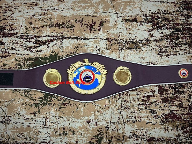 Op de afbeelding: Een gouden en blauwe bokskampioensgordel met het logo van de World Boxing Organization (WBO). De gordel heeft een bruin leren band met witte piping en twee gouden zijplaten.