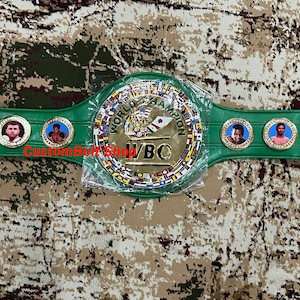 Op de afbeelding: Een groene en gouden bokskampioensgordel met het WBO-logo en meerdere vlaggen. De gordel heeft een middelste plaat met de woorden "World Boxing Organization" en "WBO" in gouden letters.