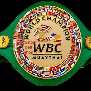 May include: A green and gold World Champion boxing belt. The belt features a large gold centre plate with the text "WORLD CHAMPION" and "WBC MUAYTHAI". The belt is adorned with flags and two smaller side plates with a figure. The text "Custom belt shop" is also visible.