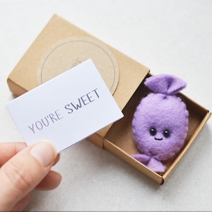 May include: A small, purple felt candy character with a smiling face sits inside a brown box. A card held nearby reads "YOU'RE SWEET". The Etsy shop's URL is visible at the bottom.