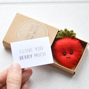 May include: A felt strawberry with a smiling face and green leaves, presented in a small cardboard box. A card with the text "I LOVE YOU BERRY MUCH" is held nearby. The box has a circular sticker with text.