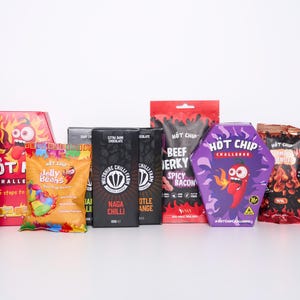 Op de afbeelding: Een verzameling voedingsproducten, waaronder een pot Volcano-ketchup, Hot Chip Challenge-dozen, Jelly Beans, pure chocoladerepen, beef jerky en Hot Chip Challenge-verpakkingen. De producten staan op een witte ondergrond.
