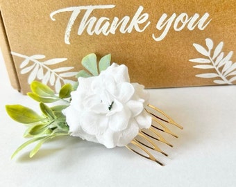 Puede incluir: Un peine floral blanco con dientes dorados, acentuado por hojas verdes y verde claro. El peine está colocado frente a una caja marrón con las palabras "Thank you" en escritura blanca.