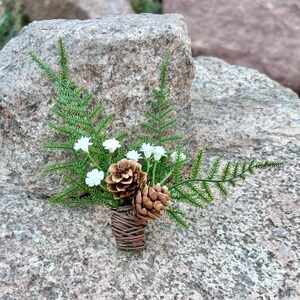 May include: A small bouquet of white flowers and pine cones wrapped in brown twine. The bouquet is sitting on a gray rock.