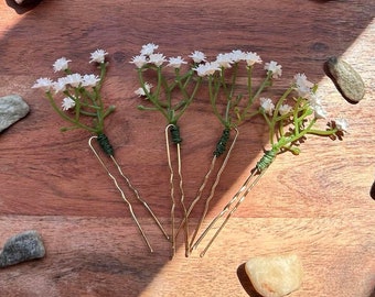Puede incluir: Cinco horquillas doradas con detalles de flores blancas. Las horquillas están dispuestas sobre una superficie de madera con pequeñas piedras esparcidas a su alrededor.