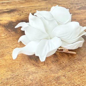 Puede incluir: Una pinza para el pelo con forma de flor blanca grande, con m&uacute;ltiples p&eacute;talos y una pinza de metal dorado. La flor es de un material suave y est&aacute; dise&ntilde;ada para el cabello. Sobre una superficie de madera.