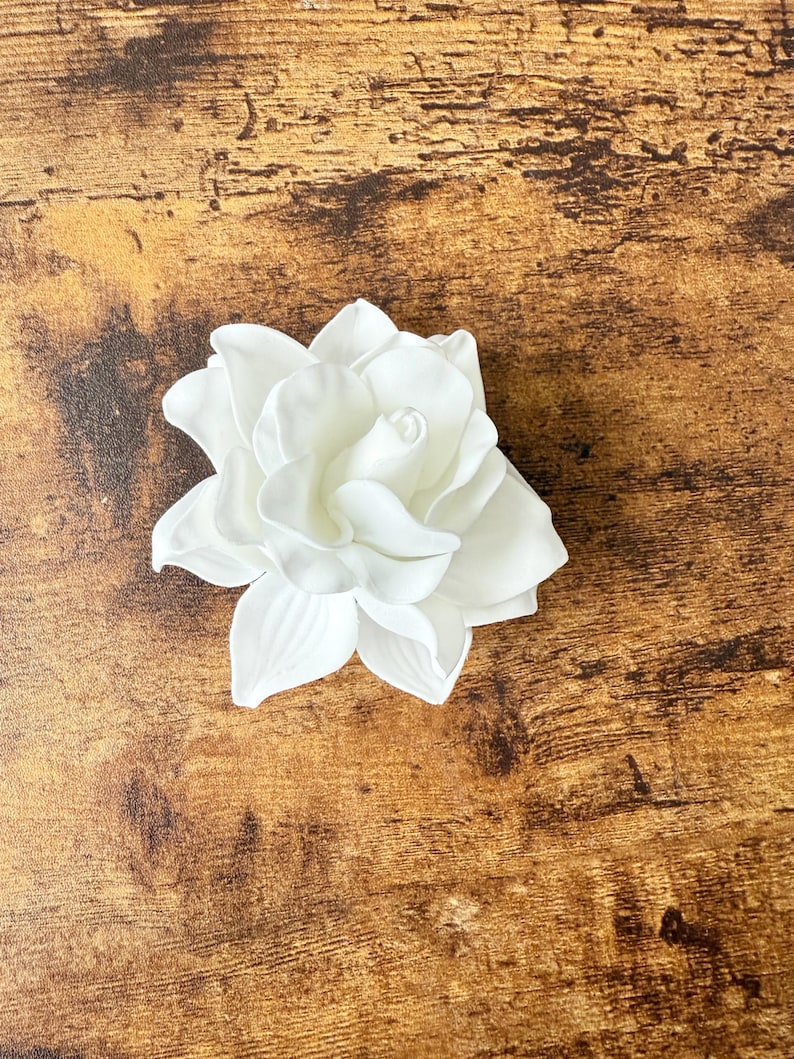 Puede incluir: Una flor de gardenia blanca artificial con m&uacute;ltiples p&eacute;talos. La flor est&aacute; en plena floraci&oacute;n y descansa sobre una superficie de madera marr&oacute;n. Los p&eacute;talos en capas crean una apariencia realista. El tema es floral y decorativo.