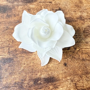 Puede incluir: Una flor artificial grande y blanca con m&uacute;ltiples capas de p&eacute;talos. La flor est&aacute; centrada y tiene una apariencia realista, con un centro en espiral y p&eacute;talos texturizados. La flor est&aacute; sobre un fondo de madera marr&oacute;n.