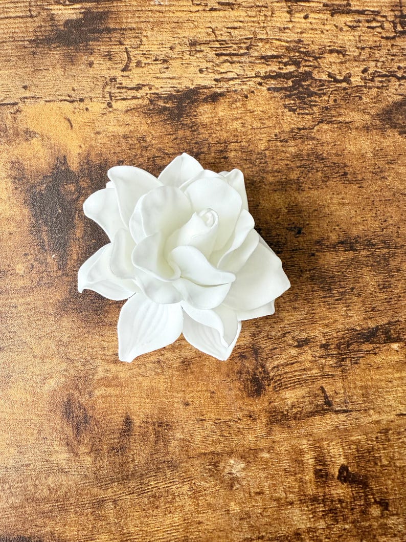 Puede incluir: Una flor de gardenia blanca artificial con m&uacute;ltiples p&eacute;talos. La flor est&aacute; en plena floraci&oacute;n, mostrando su dise&ntilde;o en capas. El fondo es una superficie de madera con vetas naturales.