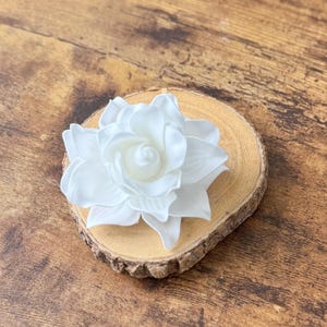 Puede incluir: Una flor artificial grande y blanca con m&uacute;ltiples p&eacute;talos, que descansa sobre una peque&ntilde;a rodaja de madera redonda. La flor est&aacute; centrada en la madera, que tiene un borde de corteza natural. El fondo es una superficie de madera.