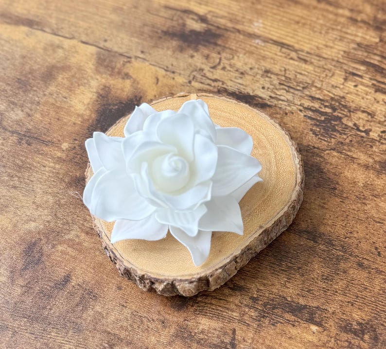 Puede incluir: Una flor artificial grande y blanca con m&uacute;ltiples p&eacute;talos, que descansa sobre una peque&ntilde;a rodaja de madera redonda. La flor est&aacute; centrada en la madera, que tiene un borde de corteza natural. El fondo es una superficie de madera.