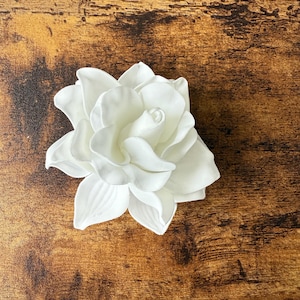Puede incluir: Una flor de gardenia blanca artificial con m&uacute;ltiples capas de p&eacute;talos. La flor est&aacute; en plena floraci&oacute;n, mostrando su delicada textura y forma. La flor est&aacute; sobre una superficie de madera marr&oacute;n.