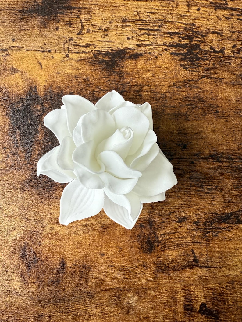 Puede incluir: Una flor de gardenia blanca artificial con m&uacute;ltiples capas de p&eacute;talos. La flor est&aacute; en plena floraci&oacute;n, mostrando su delicada textura y forma. La flor est&aacute; sobre una superficie de madera marr&oacute;n.