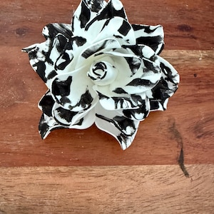 Puede incluir: Una flor decorativa hecha a mano con un patr&oacute;n en blanco y negro. La flor tiene m&uacute;ltiples p&eacute;talos con un dise&ntilde;o moteado en blanco y negro. El centro de la flor es blanco. La flor est&aacute; sobre una superficie de madera.