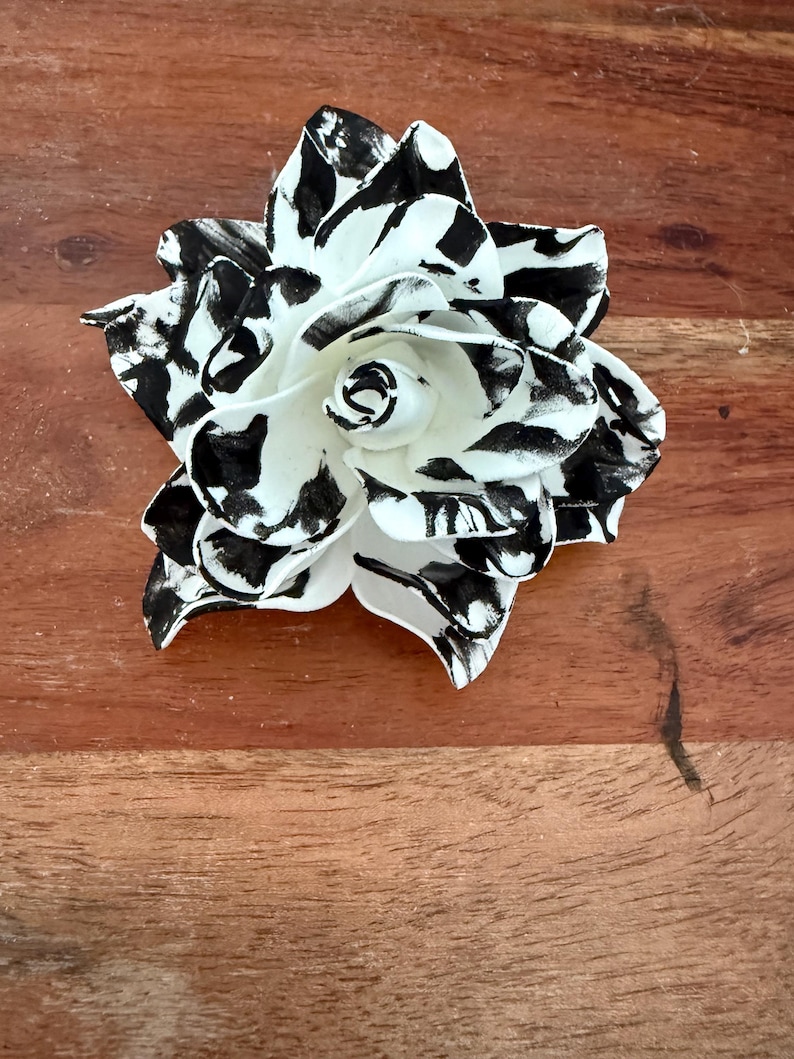 Puede incluir: Una flor decorativa hecha a mano con un patr&oacute;n en blanco y negro. La flor tiene m&uacute;ltiples p&eacute;talos con un dise&ntilde;o moteado en blanco y negro. El centro de la flor es blanco. La flor est&aacute; sobre una superficie de madera.