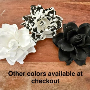 Puede incluir: Tres decoraciones de flores artificiales en blanco, negro y blanco y negro. La flor blanca tiene m&uacute;ltiples capas de p&eacute;talos. La flor en blanco y negro tiene un patr&oacute;n moteado. La flor negra tiene un dise&ntilde;o de p&eacute;talos en capas. El texto en la parte inferior dice "Otros colores disponibles al finalizar la compra."