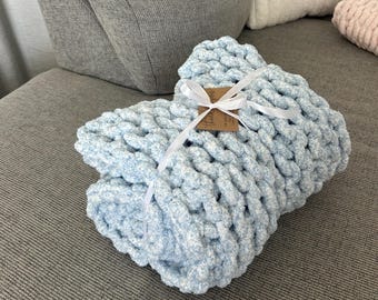 May include: A folded, chunky knit blanket in a soft, light blue colour, tied with a white ribbon. The blanket is made of thick yarn, creating a textured appearance. A small tag is attached to the ribbon.