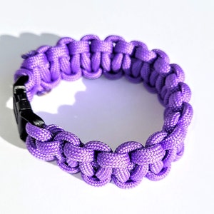 May include: A purple paracord bracelet with a black plastic buckle closure. The bracelet is made with a woven knot pattern.