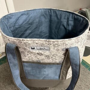 May include: A gray and white floral tote bag with a blue lining and brown straps. The bag has a blue pocket on the front and a label that reads "Going to Grandma's House".