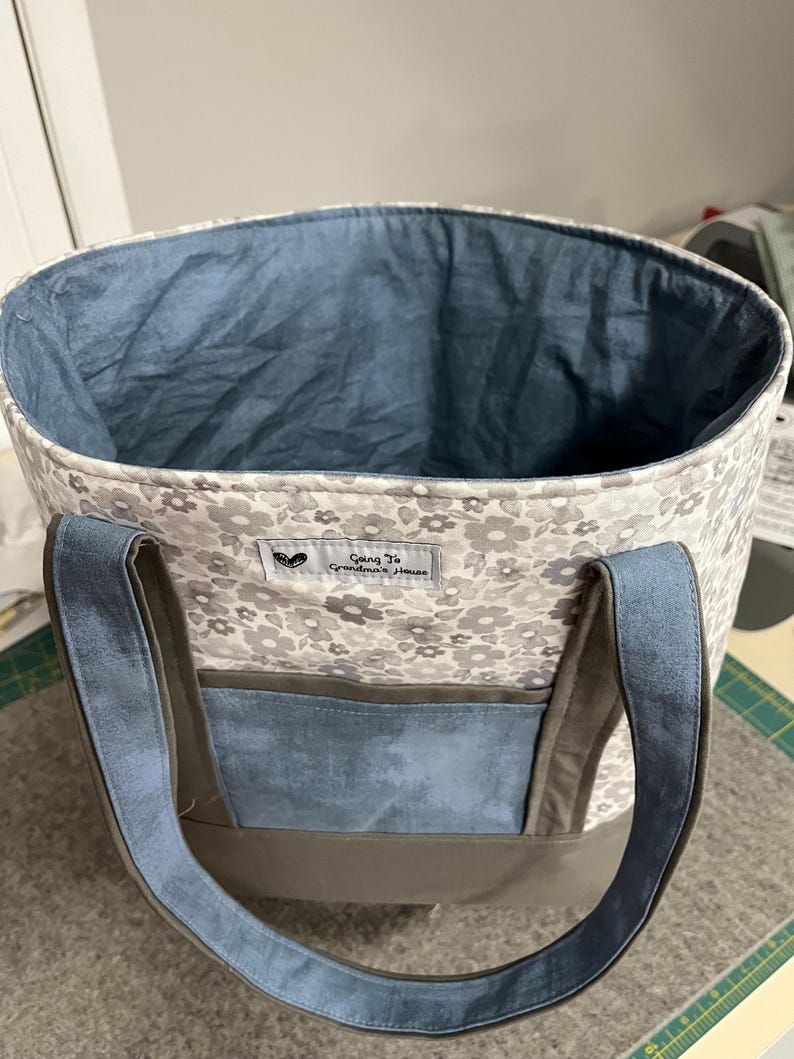 May include: A gray and white floral tote bag with a blue lining and brown straps. The bag has a blue pocket on the front and a label that reads "Going to Grandma's House".