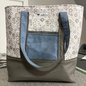 May include: A gray and white floral tote bag with blue and gray fabric accents. The bag has two long straps and a pocket on the front. The bag has a label that says "Going to Grandma's House".