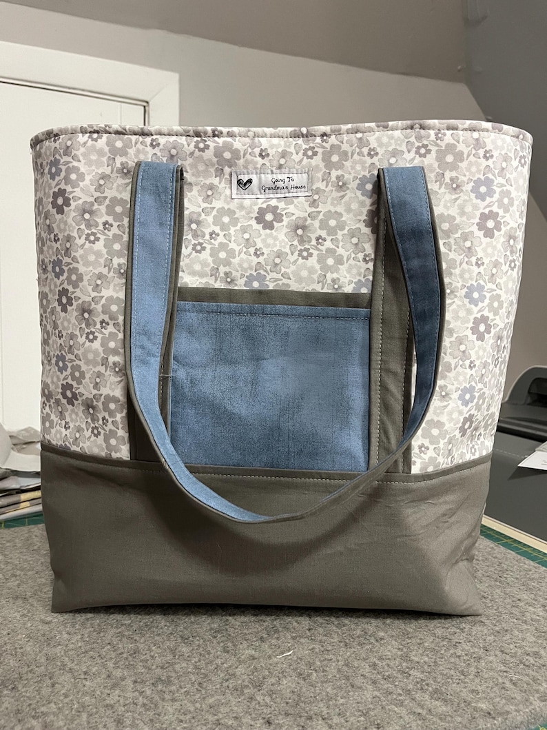 May include: A gray and white floral tote bag with blue and gray fabric accents. The bag has two long straps and a pocket on the front. The bag has a label that says "Going to Grandma's House".