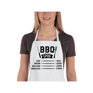 Puede incluir: Delantal blanco con texto negro que dice "BBQ Timer" y una lista de niveles de cocción para la carne con el número de cervezas correspondiente. La lista incluye: Poco hecho, Medio, Bien hecho, Pedir pizza. 1 cerveza, 2 cervezas, 3 cervezas, 6 cervezas.