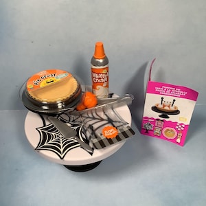 May include: A miniature Halloween-themed display featuring a pumpkin pie, whipped cream, and pie crust. The pie sits on a white cake stand decorated with a black spiderweb design. A small box reads "Pumpkin Pie".