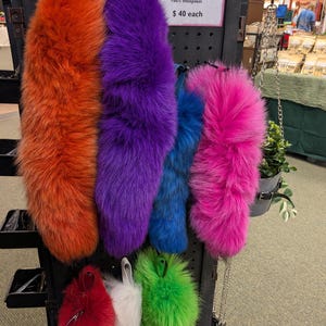 May include: A display of colorful clip-on tails in various shades, including orange, purple, blue, and pink. The tails are made of sheepskin and have a built-in snap clip attachment. The sign reads "Clip-On Tails!"