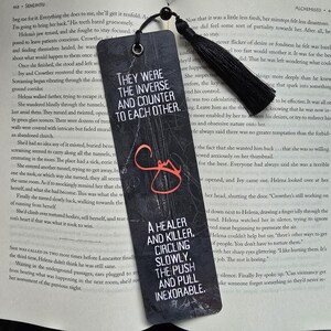 May include: Black bookmark with text: "THEY WERE THE INVERSE AND COUNTER TO EACH OTHER." and "A HEALER AND KILLER. CIRCLING SLOWLY. THE PUSH AND PULL INEXORABLE." Features a black tassel and a hole for threading.