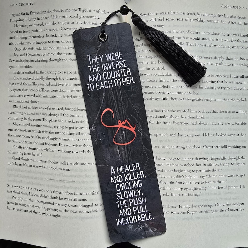 May include: Black bookmark with text: "THEY WERE THE INVERSE AND COUNTER TO EACH OTHER." and "A HEALER AND KILLER. CIRCLING SLOWLY. THE PUSH AND PULL INEXORABLE." Features a black tassel and a hole for threading.