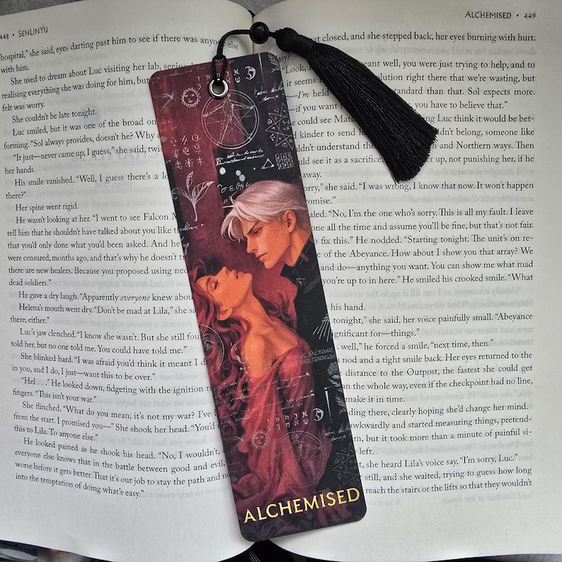 May include: A bookmark with a romantic illustration of a couple embracing, with the word "ALCHEMISED" at the bottom. The bookmark has a black tassel and is set against a book page.
