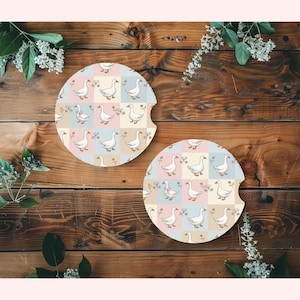 May include: Two round car coasters with a whimsical goose pattern. Each coaster features a repeating design of white geese, flowers, and butterflies on a pastel patchwork background of pink, blue, and beige squares. The coasters are designed to fit in a car's cup holder.