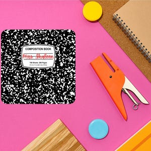Op de afbeelding: Een compositieboek met een zwart-witte gespikkelde kaft, met de tekst "COMPOSITION BOOK" en "Mrs. Shaleen" in het rood. Het notitieboek is 20,3 cm x 17,8 cm. Een oranje nietmachine, een gele knop, een blauwe knop en een spiraalgebonden notitieboek zijn ook zichtbaar.