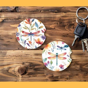 May include: Two white car coasters with watercolor dragonfly and floral designs. The coasters feature a colorful dragonfly with orange, red, and blue wings, surrounded by flowers and leaves. The coasters are on a wooden surface with car keys.