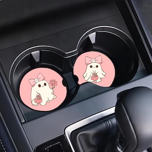 May include: Two pink car coasters with a cartoon ghost design. Each coaster features a white ghost with a pink bow, holding a flower and a basket. The coasters are placed in a car's cup holders.