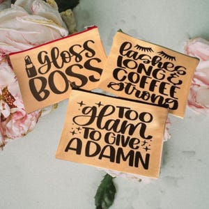 Könnte beinhalten: Drei beigefarbene Reißverschlusstaschen mit schwarzem Text. Die Taschen haben folgenden Text: "gloss boss", "lashes long & coffee strong" und "too glam to give a damn".