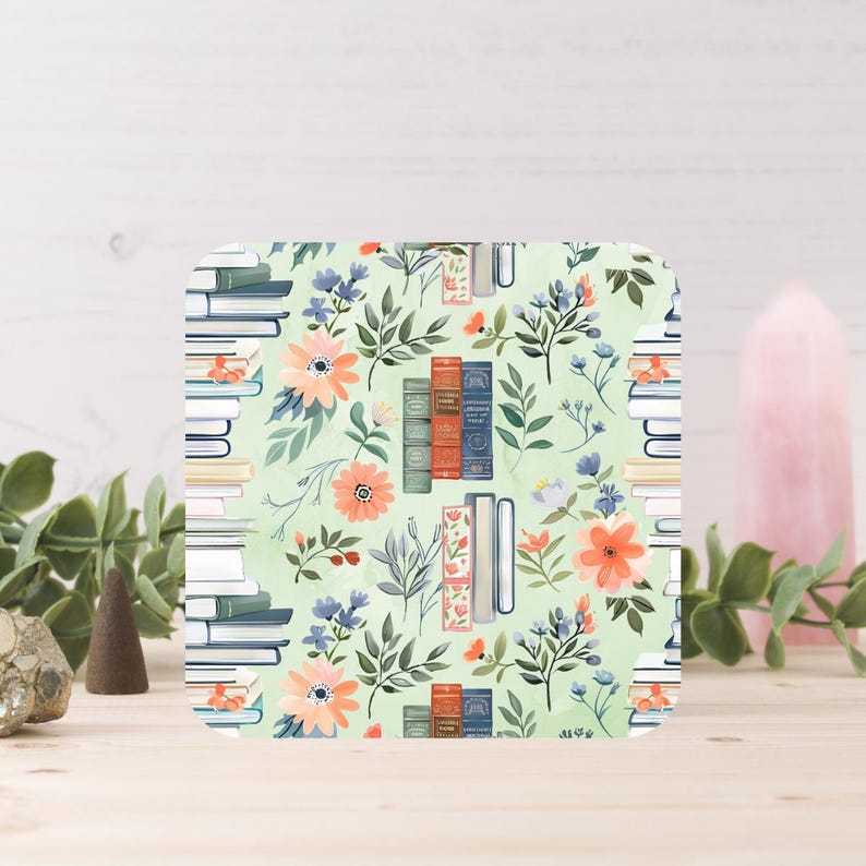 May include: Square coaster with a floral and book design. The coaster features stacks of books, orange and blue flowers, and green foliage on a light green background. The coaster is made of a durable material.
