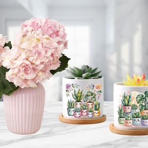 May include: A pink vase with a bouquet of pink hydrangeas sits next to two white ceramic planters with illustrations of potted plants. The planters are on wooden saucers. One planter has a succulent, the other has a yellow flower.