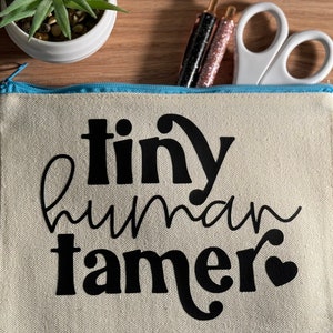 May include: A white canvas zippered pouch with a blue zipper. The pouch has black text that reads "tiny human tamer" with a small heart.
