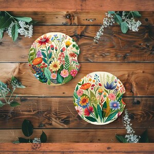 May include: Two round car coasters with a floral design. The coasters feature a variety of colorful flowers, including red, pink, yellow, and blue blooms, set against a cream background. The coasters are displayed on a wooden surface.