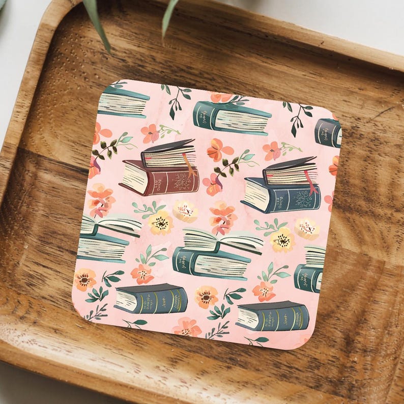 May include: Square coaster with a pink background featuring a pattern of books and floral elements in shades of brown, blue, and orange. The books are in various positions, some open, some closed.