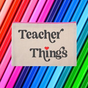 May include: A beige canvas pencil case with a pink zipper. The case has the text "Teacher Things" in black with a red heart between the words.