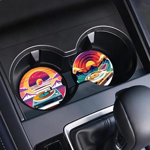 May include: Two car coasters with a retro sunset and turntable design. The coasters feature a vibrant color palette of orange, purple, and teal. The coasters are placed in the car's cup holders, adding a touch of vintage flair to the vehicle's interior.